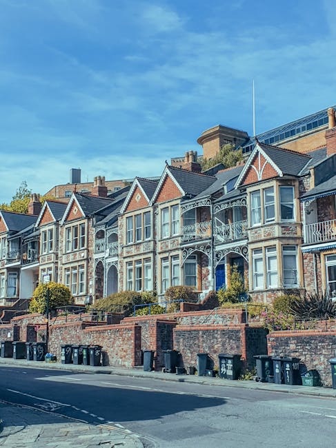 A row of Victorian-style terraced houses situated on a residential street, featuring decorative cast-iron balconies, bay windows, and gabled roofs with red brick and white trim. The buildings are set 