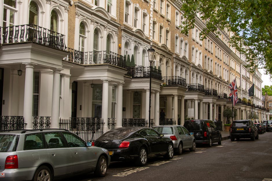A row of Victorian-style terraced houses situated on a residential street, featuring decorative cast-iron balconies, bay windows, and gabled roofs with red brick and white trim. The buildings are set 