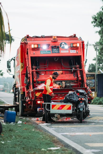 A sanitation worker dressed in a yellow vest and black trousers stands behind a large red rubbish collection truck, which is positioned on the side of a street. The back of the truck is open, revealin