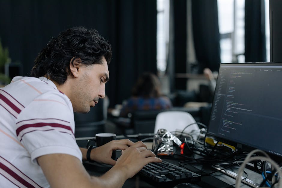 A young man with dark, slightly curly hair and light skin is seated at a desk in an office or co-working space, focusing on typing on a black keyboard. He is wearing a white short-sleeved shirt with r